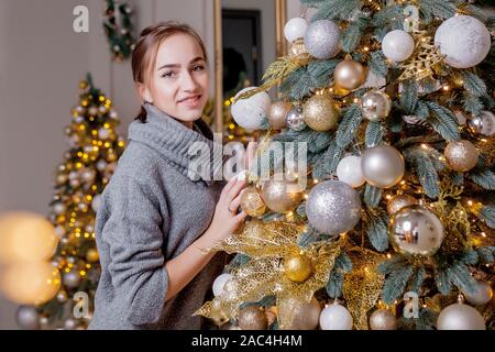 Bella ragazza decorare albero di Natale. Una giovane donna sorridente prepara un albero di Natale per la vacanza. Lussureggiante verde albero di Natale con palline dorate Foto Stock