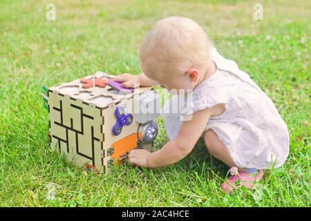 Carino bambina sta giocando con busiboard all'aperto sull'erba verde. Giocattoli educativi per i bimbi. ragazza porta aperta a cubo del bordo Foto Stock