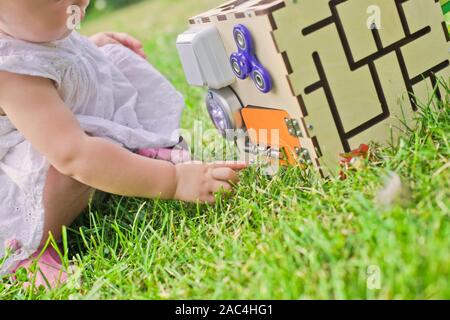 Carino bambina sta giocando con busiboard all'aperto sull'erba verde. Giocattoli educativi per i bimbi. ragazza porta aperta a cubo del bordo Foto Stock