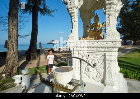 Un giovane ragazzo appare in un santuario con un quattro-di fronte Brahma (dio indù) statua nel parco di Dusit Laguna Hotel, Bang Tao Beach, Phuket, Tailandia Foto Stock