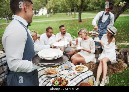 I camerieri servono gruppo di buoni amici che siedono fuori sul bellissimo campo con alberi Foto Stock