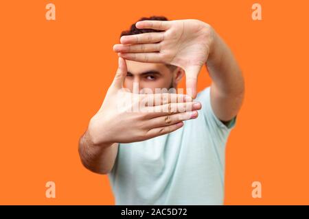 Ritratto di attenti curiosi brunette uomo in casuale t-shirt bianco guardando la fotocamera con un occhio, mettere a fuoco attraverso il photo frame fatto di dita. indoo Foto Stock