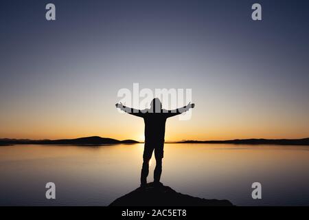 Uomo di silhouette sta con le braccia sollevate contro il tramonto sul lago e sui monti Foto Stock