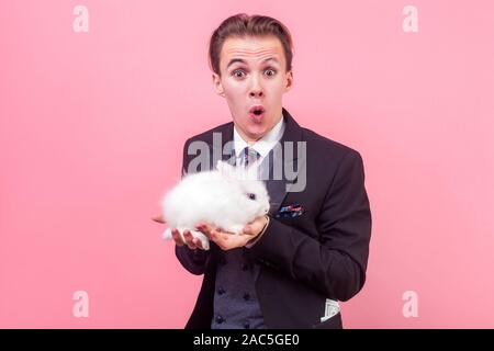 Adozione di Pet. Ritratto di estremamente entusiasti uomo in elegante abito elegante con taglio di capelli tenendo simpatico coniglio sul suo palm, guardando la fotocamera con stupore Foto Stock