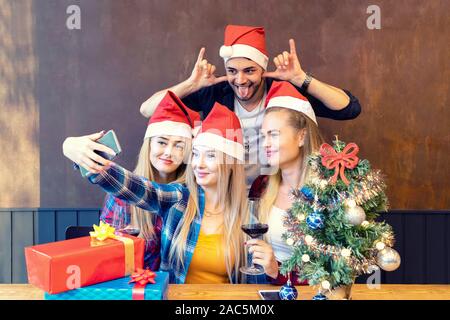 Felice giovani amici indossando santa claus hat godendo di Christmas party tenuto selfie con facce buffe a casa da condividere sui social media Foto Stock