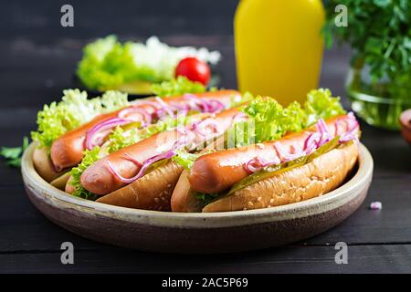 Hot Dog con salsiccia, cetriolo sottaceto, pomodori, cipolle rosse e lattuga su legno scuro dello sfondo. Estate hotdog. Foto Stock