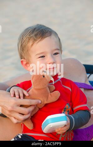 Un one-anno-vecchia parte-Asian boy a Big Island Beach detiene un tricheco farcite giocattolo con sua madre aiutare. Foto Stock