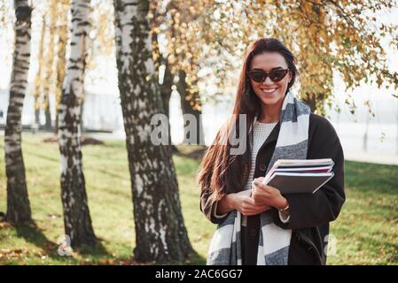 Giovani sorridente brunette in occhiali da sole sorge nel parco vicino agli alberi e mantiene il blocco note Foto Stock