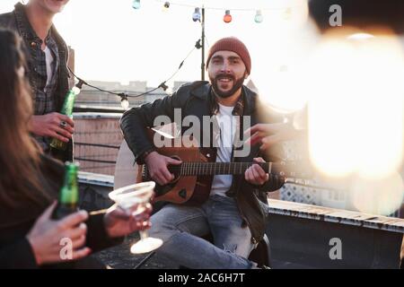 Chitarrista barbuto sorride alla telecamera. Tre amici con alcool e la chitarra acustica. Lampadina sfocata nel lato destro della foto Foto Stock