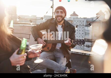 Felice bello ragazzo con la chitarra acustica sorride alla telecamera e si siede sul tetto con i suoi amici Foto Stock