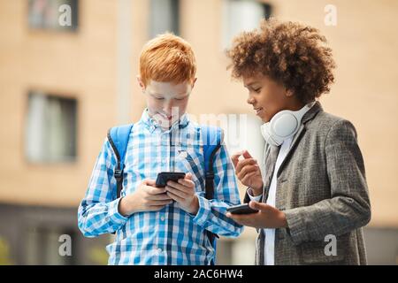 Due scolari in piedi con i telefoni cellulari a discutere di qualcosa e studiare online all'aperto Foto Stock