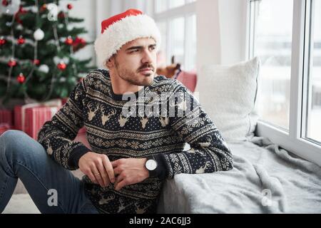 Appoggiata sul davanzale della finestra. Foto di un uomo in santa hat e vacanze vestiti guarda attraverso la finestra. Albero di natale sullo sfondo Foto Stock