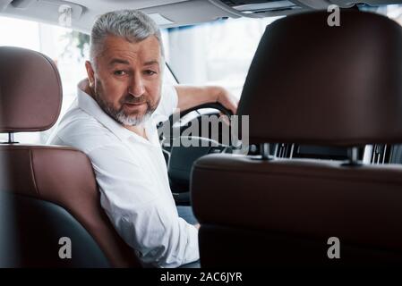 Gioioso uomo barbuto in bianco camicia guarda verso la fotocamera mentre è seduto in una macchina moderna Foto Stock