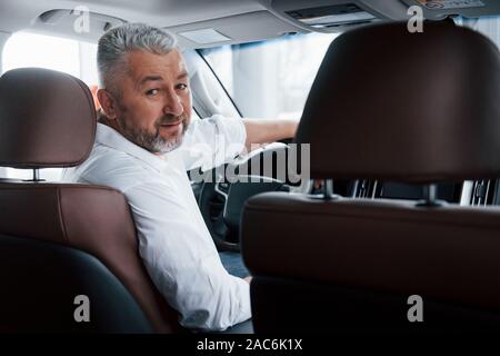 Concezione di successo. Gioioso uomo barbuto in bianco camicia guarda verso la fotocamera mentre è seduto in una macchina moderna Foto Stock