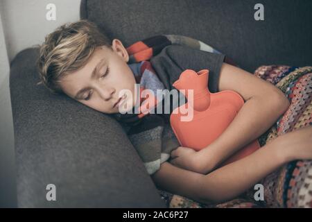 Ragazzo malato a letto con coperta di lana e bottiglie di acqua calda. Triste teen con l'influenza si appoggia a casa da solo in una fredda giornata invernale. Bambino con infettare stagionali Foto Stock