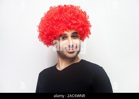 Ritratto di divertenti tanga elegante uomo con setole e ricci parrucca rosso sulla sua testa sorridente alla fotocamera, cercando giocoso positivo, ottimista lifestyle. in Foto Stock