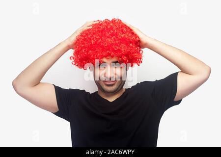 Ritratto di crazy hipster, gioiosa uomo con setole e in t-shirt toccando parrucca rosso sulla sua testa e sorridente alla fotocamera, cercando divertente con capelli luminosi. Foto Stock