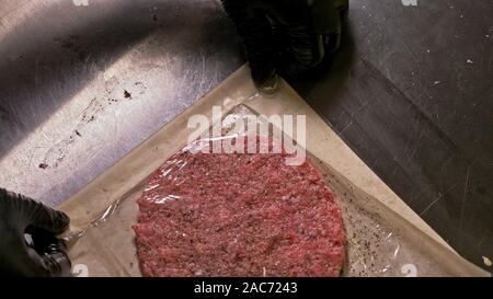 Cuocere rende Medaglioni di carne per gli hamburger. Chef in nero guanti di cibo rende cotoletta. Cotolette siano livellati in acciaio anello in ancora un medaglione. Sulla parte superiore di versare Foto Stock