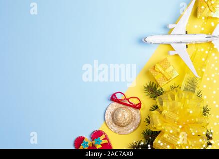 Natale concetto di viaggio. Rami di abete con bowknot e neve, confezioni regalo, giocattolo flop, cappello, occhiali da sole e aereo su giallo-blu sullo sfondo. Vista dall'alto, Foto Stock