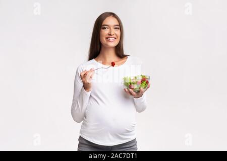 Sorridente donna incinta di mangiare insalata di verdure in piedi, Studio Shot Foto Stock