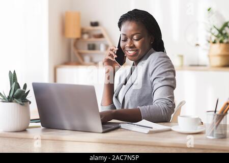 Imprenditrice Afro parlando al cellulare e utilizzando computer portatile in ufficio Foto Stock