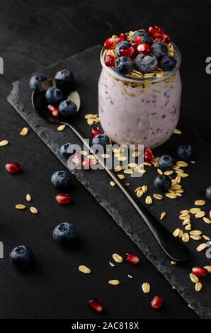 Colazione sana nozione. Vasetti di yogurt fatto in casa con mirtilli, i semi di melograno e avena su sfondo nero. Verticale. Foto Stock