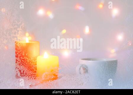 Rosso e giallo candele su sfondo bianco. Tazza di caffè all'interno della casa con la brina sul vetro e luci di noi un simbolo di vacanza. Foto Stock