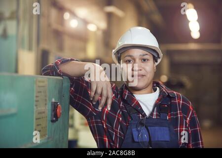Ritratto del moderno lavoratore di sesso femminile che sorride alla telecamera mentre appoggiata sulle unità della macchina in stabilimento industriale, spazio di copia Foto Stock