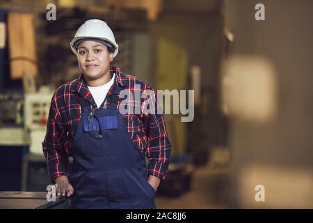 Vita su ritratto del moderno lavoratore di sesso femminile che guardando la fotocamera mentre appoggiata sulle unità della macchina in stabilimento industriale, spazio di copia Foto Stock