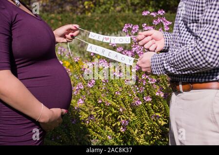 La gravidanza di una coppia ottiene pronto a diventare una famiglia in un matnerity photoshoot dove tengono un simpatico segno per il bambino Foto Stock