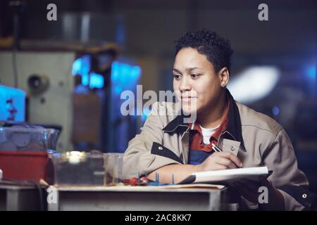 Ritratto di sorridere donna afro-americana a lavorare alla linea di produzione in fabbrica e prendere appunti su appunti, spazio copia Foto Stock