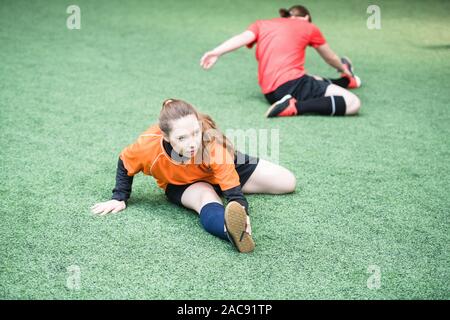 Piccolo gruppo di attivi calciatori femmina in sport uniforme di fare esercizio per distendere le gambe mentre è seduto sul campo Foto Stock