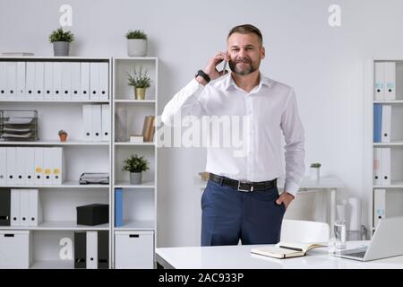 Successo imprenditore maturo nel convenzionale parlare su smartphone al cliente o partner aziendali in ufficio Foto Stock