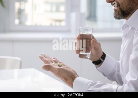 Le mani del barbuto imprenditore pillola di contenimento mentre stava andando a prendere e bicchiere di acqua sulla scrivania a lavoro Foto Stock