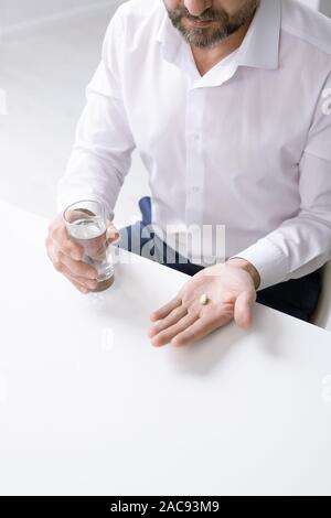 Le mani del barbuto imprenditore con mal di testa tenendo pillola e bicchiere di acqua sulla scrivania in ufficio Foto Stock