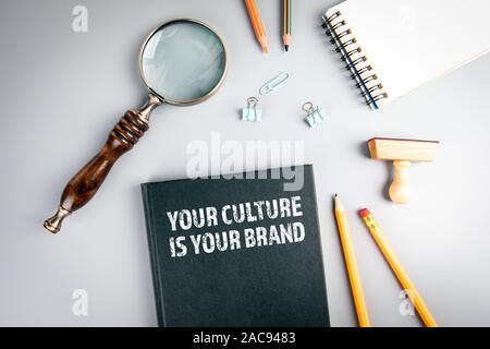 La vostra cultura è il tuo marchio. La conoscenza, le competenze e il concetto di commercializzazione. Libro nero coperchio e lente di ingrandimento su un tavolo grigio Foto Stock
