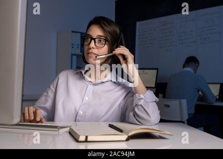Giovani femmine hotline operatore con cuffia parlando ai clienti online di fronte allo schermo del computer Foto Stock