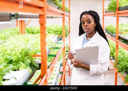I giovani africani gravi biologo femmina guardando a voi mentre si effettua la ricerca e le note durante il lavoro in serra Foto Stock