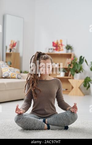 Piuttosto giovane donna attiva con dreadlocks seduto sul pavimento con le gambe incrociate Foto Stock