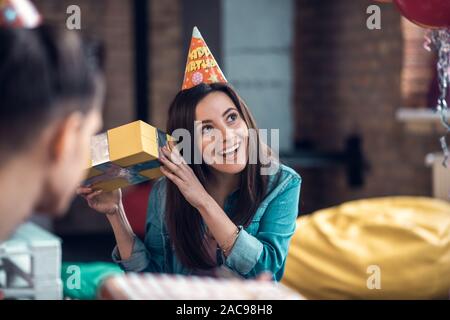 Curioso circa il presente. Dark-eyed allegro compleanno ragazza curiosa sensazione dopo aver ricevuto il regalo di compleanno Foto Stock