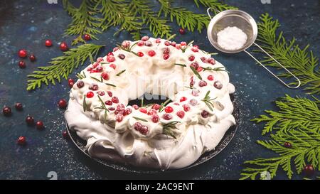 Torta di Natale Pavlova con mirtillo rosso decorato con foglie di rosmarino su sfondo blu scuro accanto a rami di abete. Foto Stock