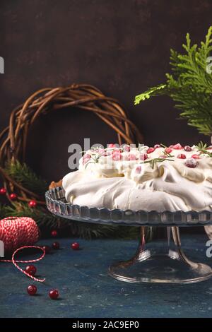 Torta di Natale Pavlova con mirtillo rosso decorato con foglie di rosmarino su sfondo blu scuro accanto a rami di abete. Foto Stock