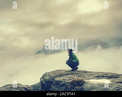 Giovane maschio viaggiatore seduto sulla scogliera di arenaria. Vista posteriore dei maschi di un escursionista in cappello e giacca verde siton il picco roccioso mentre godendo di un alba abo Foto Stock