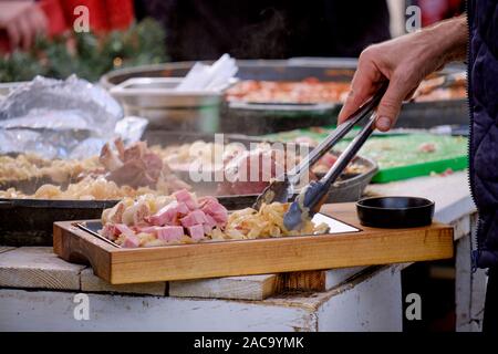 Focus sulla tradizionale croata Buncek- Hock di maiale preparato in una padella grande. Mano di uomo con le tenaglie di maiale di placcatura e il cavolo da cottura preparazione Foto Stock