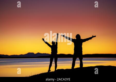 Sagome di due escursionisti si erge in vincitore posano con braccia sollevate contro il tramonto sul lago e sui monti Foto Stock