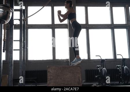 Montare la giovane donna box jumping in un stile crossfit palestra. Atleta femminile è di eseguire la casella salta in palestra Foto Stock