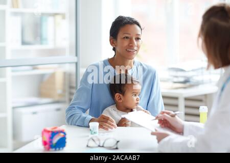 Giovane madre insieme con il suo servizio di baby sitter e parlando con il pediatra all'ufficio del medico Foto Stock