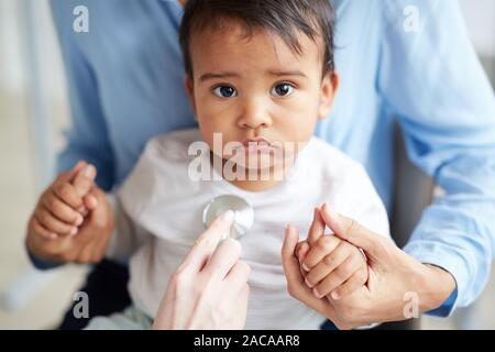 Ritratto di bambino guardando la fotocamera mentre medico esaminando lui con uno stetoscopio e madre seduta in background Foto Stock