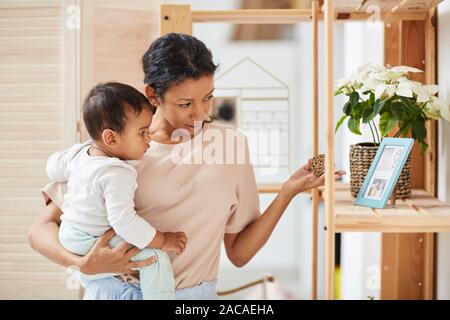 Giovane madre mentre tiene il suo bambino sulle sue mani e a mostrargli alcune interessanti oggetto dal ripiano a casa Foto Stock