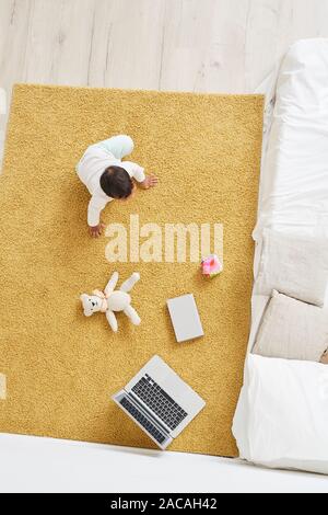 Angolo di alta vista del bambino seduto sul tappeto vicino al computer portatile e giocattoli in camera Foto Stock